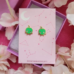 May include: A pair of green apple-shaped earrings with gold-colored trim and a small leaf on top. The earrings are displayed on a pink card with a constellation design and the text "@MAINTAININGMEDIOCRITY". The earrings are in a pink box.