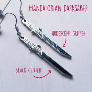 Puede incluir: Un par de pendientes de plata con forma de sable oscuro mandaloriano. Un pendiente tiene purpurina negra y el otro tiene purpurina iridiscente.