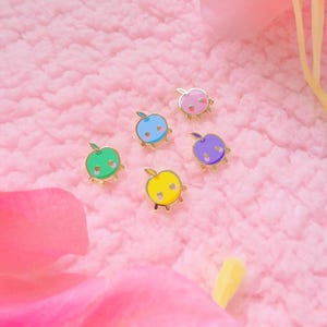 May include: Five small enamel pins shaped like apples in pink, blue, green, yellow, and purple. Each apple pin features a gold-toned metal outline, a small stem, and two circular cutouts representing eyes, creating a simple, stylized fruit character design.