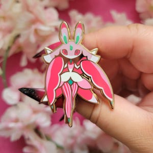 May include: A pink and gold enamel pin depicting a stylized creature with bunny ears and wing-like features. The pin has a gold outline and is set against a pink backdrop with flowers.