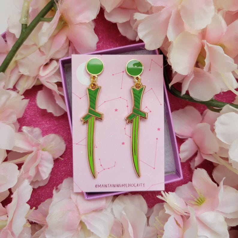 K&ouml;nnte beinhalten: Ein Paar goldfarbene Ohrringe mit einem gr&uuml;nen Dolch-Design. Jeder Ohrring hat einen gr&uuml;nen Kreis oben, einen gr&uuml;nen und goldenen Griff und eine lange, gr&uuml;ne Klinge. Die Ohrringe werden in einer rosa Schachtel mit dem Text "@MAINTAININGMEDIOCRITY" pr&auml;sentiert.