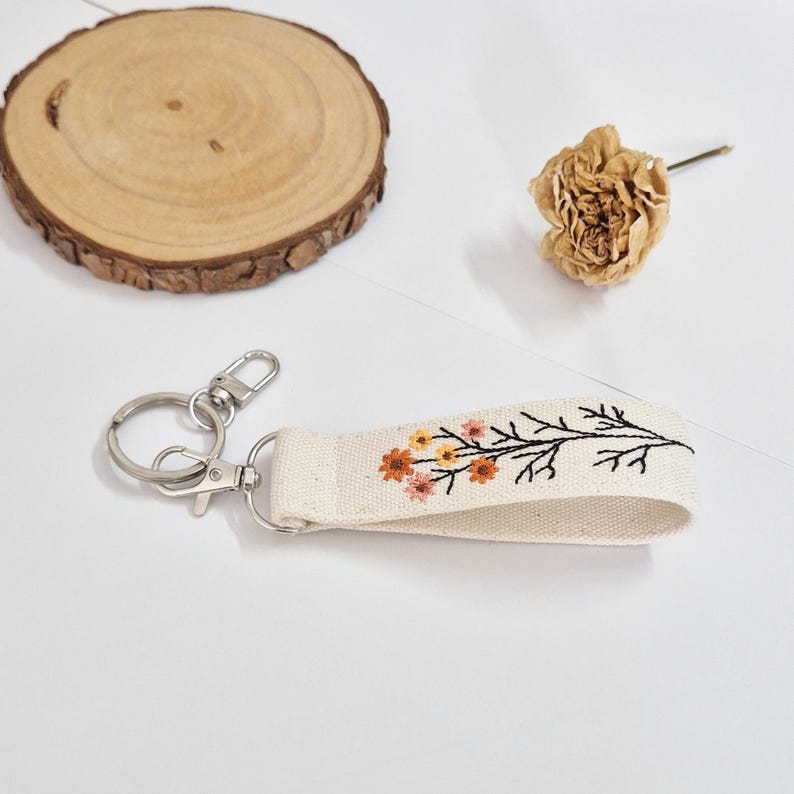 May include: A cream-colored keychain wristlet with a floral embroidery design in orange, yellow, and red. The keychain has a silver clasp and ring. A wooden slice and dried flower are in the background.