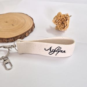 May include: A cream-colored canvas keychain with the name "Ashlynn" embroidered in black script. The keychain has a silver-colored metal clasp and ring. The background includes a wooden slice and a dried flower.