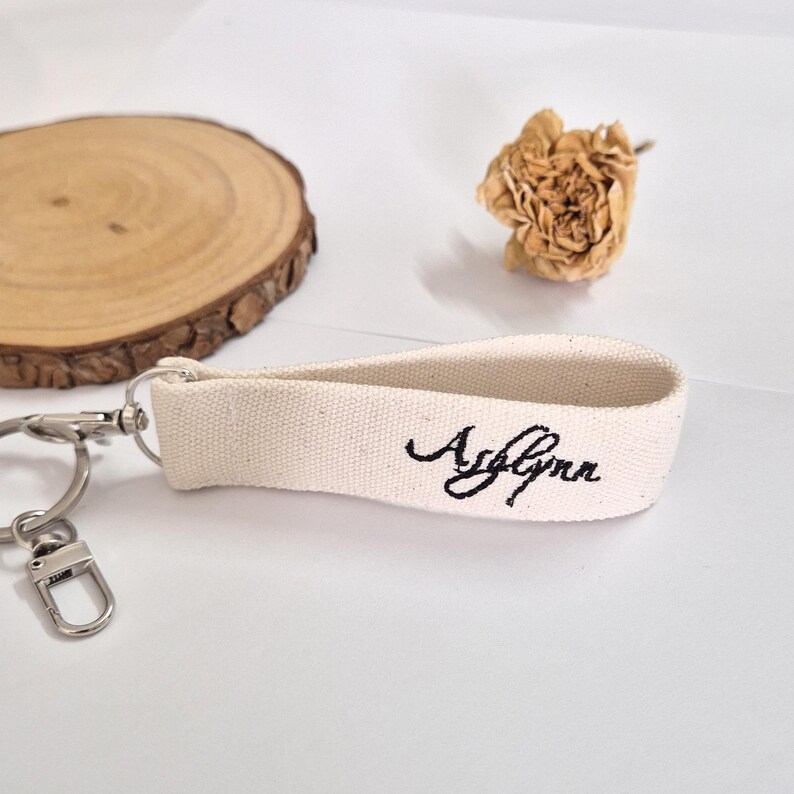 May include: A cream-colored canvas keychain with the name "Ashlynn" embroidered in black script. The keychain has a silver-colored metal clasp and ring. The background includes a wooden slice and a dried flower.