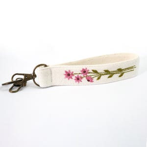 Op de afbeelding: Crèmekleurige stoffen sleutelhanger polsband met bloemenborduurwerk. De borduursels tonen roze bloemen en groene stelen. Het heeft een bronzen metalen sluiting en ring om sleutels aan te bevestigen. De lengte is ongeveer 15 cm.