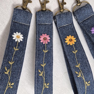 May include: Four denim fabric key fob wristlets featuring embroidered floral designs. Each strap includes a bronze-toned metal lobster clasp. The embroidery depicts individual flowers with white, pink, yellow, and purple petals on green stems.
