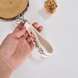 May include: Cream-colored wristlet keychain with a floral embroidery design in orange and yellow. The keychain has a silver-colored clasp. The wrist strap is made of a sturdy canvas material.