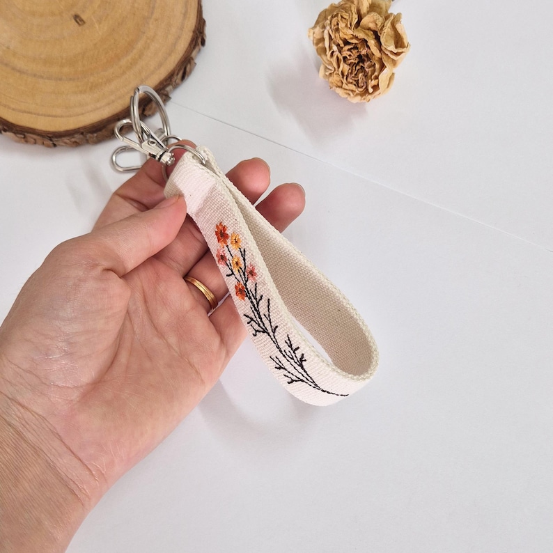 May include: Cream-colored wristlet keychain with a floral embroidery design in orange and yellow. The keychain has a silver-colored clasp. The wrist strap is made of a sturdy canvas material.