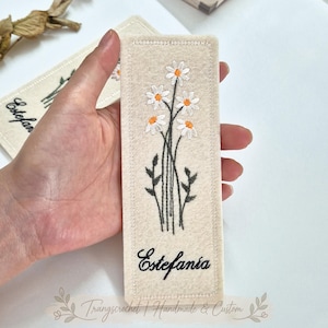 May include: A rectangular cream-coloured felt bookmark featuring an embroidered design of white daisies with orange centres and green stems. The name Estefania is embroidered in black cursive script at the bottom. The edges are finished with white stitching.
