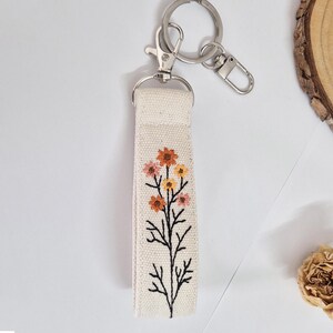 May include: A cream-colored fabric keychain strap with an embroidered floral design in orange, pink, and yellow. The strap is attached to a silver-colored metal keyring and clasp. The floral design features a black stem.