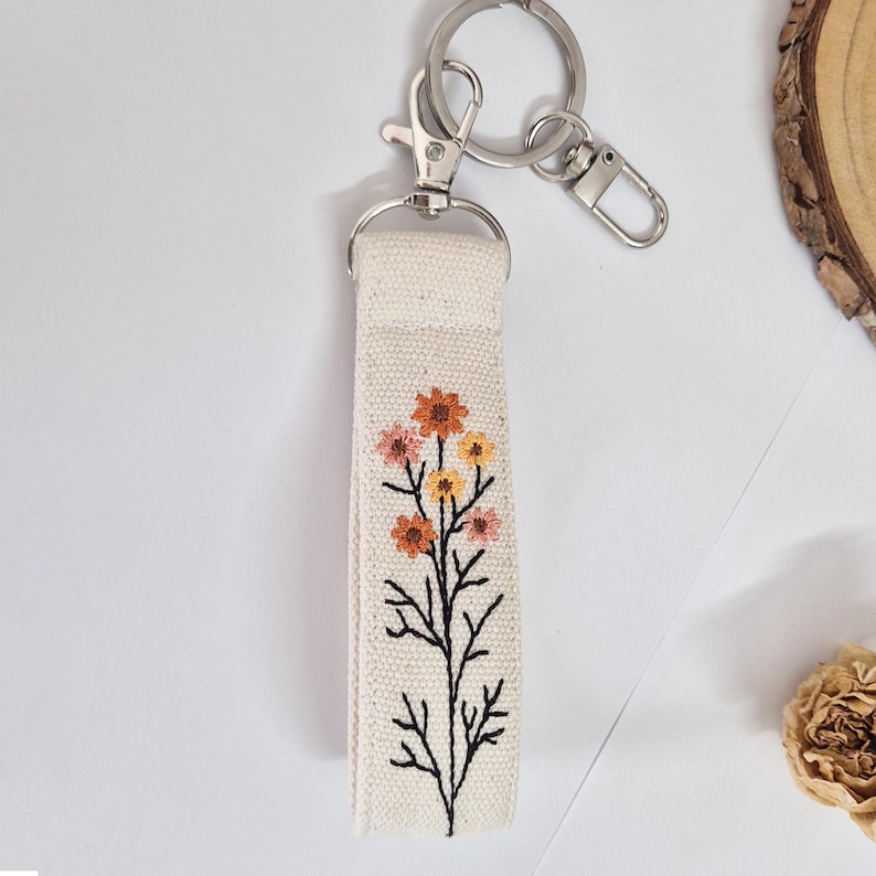 May include: A cream-colored fabric keychain strap with an embroidered floral design in orange, pink, and yellow. The strap is attached to a silver-colored metal keyring and clasp. The floral design features a black stem.