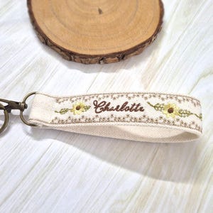 Peut inclure: Bracelet de porte-clés en tissu crème avec un motif floral brodé et le nom "Charlotte" en écriture marron. Le bracelet est doté d'un fermoir et d'un anneau dorés pour attacher les clés. L'arrière-plan comprend une tranche de bois et une surface en bois clair.