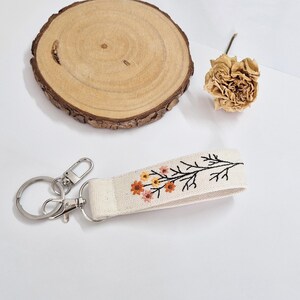 May include: A cream-colored keychain wristlet with a floral embroidery design in orange, yellow, and pink. The keychain has a silver clasp and ring. A wooden slice and dried flower are in the background.