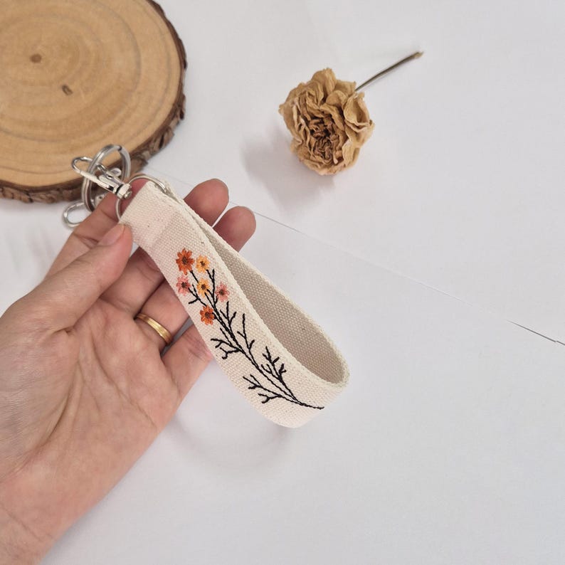 May include: A beige fabric wristlet keychain with a floral embroidery design in orange and yellow. The keychain has a silver-colored clasp. The floral design is on a black stem.