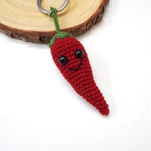 May include: A red crocheted chilli pepper keyring with a smiling face and black eyes. The keyring is attached to a silver key ring.