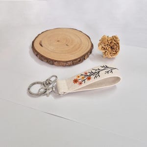 May include: A cream-colored keychain wristlet with a floral embroidery design in orange, yellow, and black. The keychain has a silver-colored clasp and key ring. A small, dried flower and a wooden coaster are also in the image.