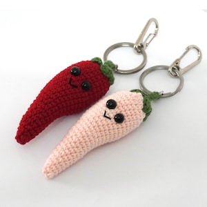 Może przedstawiać: Dwa szydełkowe breloczki do kluczy w kształcie papryki chili. Jeden jest czerwony, drugi jasnoróżowy. Oba mają czarne guziki jako oczy i uśmiechnięte twarze. Łodygi są zielone. Każdy breloczek ma srebrny metalowy pierścień i karabińczyk.