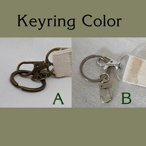 May include: Image showing two keyring options, labeled A and B. Option A features a bronze-colored clasp and ring, while option B has a silver-colored clasp and ring. Both keyrings have a beige fabric strap.
