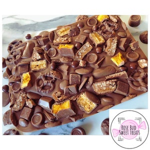 May include: A large chocolate bar with a variety of toppings, including pretzels, honeycomb, and different types of chocolate. The bar is on a marble surface and has a pink and white logo that says "Rose Bud Sweet Treats".