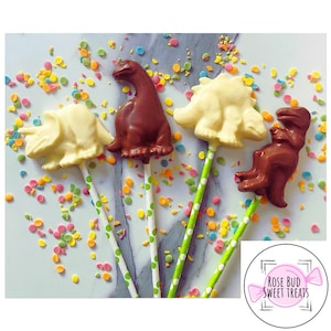 May include: Four chocolate dinosaur lollipops on green and white striped sticks. The dinosaurs are a triceratops, a stegosaurus, a brachiosaurus, and a tyrannosaurus rex. The lollipops are on a white surface with colorful sprinkles.