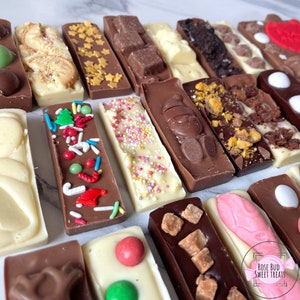 May include: An assortment of chocolate bars with various toppings, including sprinkles, stars, and candies. The bars are in different shades of brown and white chocolate, arranged on a light-colored surface. Some bars have holiday-themed decorations.