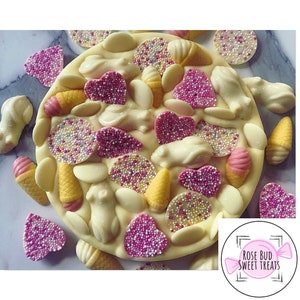 May include: White chocolate bark with pink and yellow sprinkles, hearts, ice cream cones, and bunny shapes. The bark is surrounded by additional sprinkles, hearts, and cones. The bark has the text "Rose Bud Sweet Treats" on it.