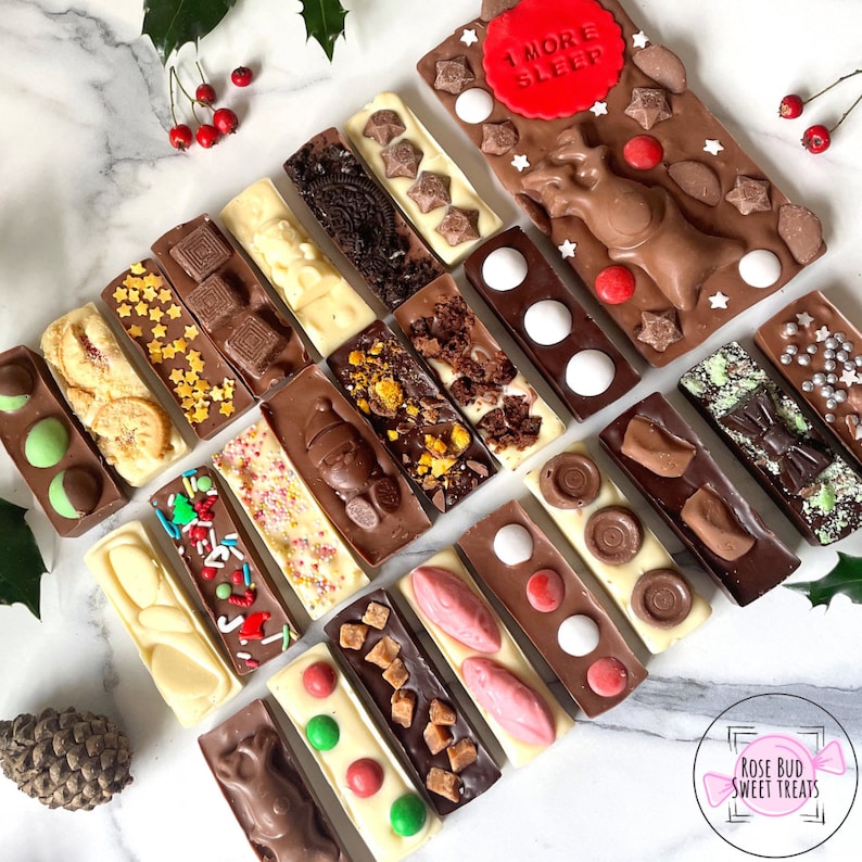 May include: An assortment of decorative chocolate bars with various toppings, including cookies, sprinkles, and candies. Some bars feature festive designs like stars and a reindeer, with one bar reading "1 MORE SLEEP". The chocolates are arranged on a marble surface.