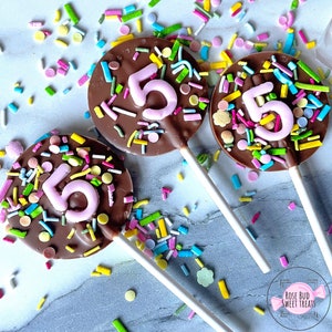 May include: Three chocolate lollipops decorated with colorful sprinkles and a pink number "5" on each. The lollipops are on a gray surface with more sprinkles scattered around.