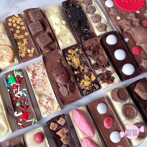 May include: An assortment of chocolate bars with various toppings, including stars, sprinkles, cookies, and candies. The bars are in milk, dark, and white chocolate. Some bars have festive decorations, and one has the word "SLEEP" on it.