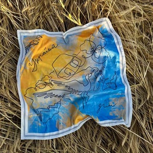 Puede incluir: Un pañuelo cuadrado de seda con un diseño de mapa en azul y amarillo, con la palabra "Україна" escrita en negro. El pañuelo tiene un borde blanco y está sobre una cama de trigo.