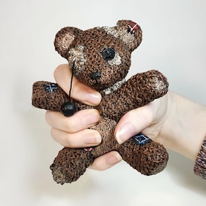 May include: A brown and black patchwork teddy bear with a black button nose and a black string with a black bead hanging from its neck.