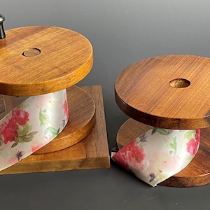 Puede incluir: Dos dispensadores de cinta de madera con cinta floral. Los dispensadores están hechos de madera de tonos cálidos y tienen una parte superior e inferior circulares. Un dispensador tiene una base cuadrada y un pomo negro. La cinta presenta flores rojas y rosas.