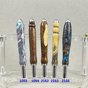 May include: Five handcrafted pen sets with unique wooden and marbled resin barrels. The pens are displayed in a clear acrylic stand. Each pen has a silver-colored tip and a different color and pattern. The numbers 1093, 1094, 2162, 2163, and 2166 are visible.