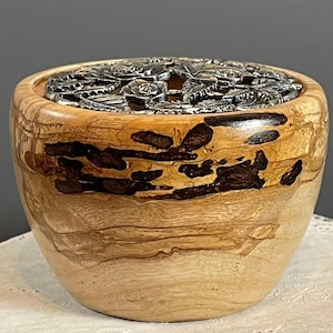 Puede incluir: Un cuenco de madera con una tapa decorativa de metal plateado. El cuenco está hecho de madera clara con nudos y vetas de color marrón oscuro. La tapa presenta un diseño floral. El cuenco es redondo y se apoya sobre una superficie blanca.