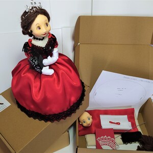 Könnte beinhalten: Eine Puppe mit einem roten Satin-Kleid und schwarzer Spitzenborte, die eine Krone trägt, sitzt in einem Karton. Die Schachtel enthält einen Puppenkopf, Stoff und andere Bastelmaterialien.