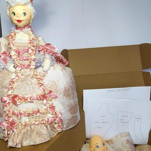 May include: A doll kit with a doll head, fabric, feathers, and a pattern sheet. The doll is wearing a pink and white dress with a lace trim. The pattern sheet has the text "PATRONES MARIA ANTONIETA" on it.