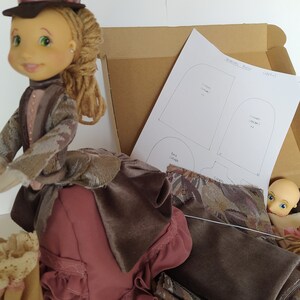Könnte beinhalten: Ein Puppen-Bastelset mit einer Stoffpuppe, Stoffresten und einem Schnittmusterbogen. Der Schnittmusterbogen enthält Anleitungen zum Nähen eines Kleides, eines Rocks und von Ärmeln. Die Puppe trägt ein braunes und rosafarbenes Kleid mit einem braunen Hut.