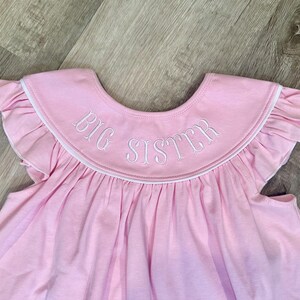 May include: A light pink baby dress with a ruffled collar and sleeves. The collar features the embroidered text "BIG SISTER" in white. The dress is made of a soft fabric.