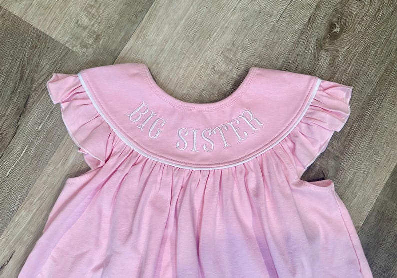 May include: A light pink baby dress with a ruffled collar and sleeves. The collar features the embroidered text "BIG SISTER" in white. The dress is made of a soft fabric.