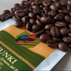 May include: A pile of dark roasted coffee beans resting on a white bag with a green and orange label. The label features a graphic of a bird and the text TUNKI, Quechua y Aymara del P, and GOURMET with five stars printed in gold.