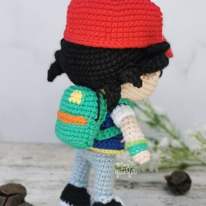 May include: Handmade crocheted doll of a character wearing a red cap, black hair, and a green backpack with an orange accent. The doll is wearing a blue and white striped shirt, light blue trousers, and black shoes.
