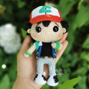 May include: Handmade crochet doll of a character wearing a white and red cap with a green letter, black shirt, blue vest, and light blue trousers. The doll has large black eyes and is held in a hand. A handcrafted item.