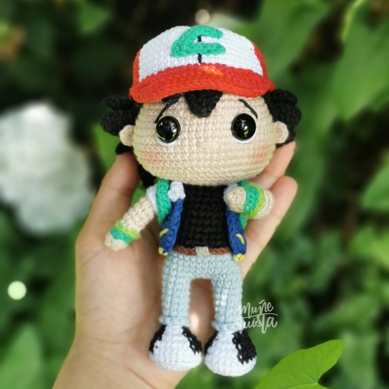 May include: Handmade crochet doll of a character wearing a white and red cap with a green letter, black shirt, blue vest, and light blue trousers. The doll has large black eyes and is held in a hand. A handcrafted item.