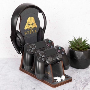 Può includere: Un supporto in legno con un design nero di Darth Vader, il nome "STEVE" stampato sotto, contiene un paio di cuffie nere e due controller per videogiochi neri. Una piccola pianta grassa in vaso si trova a destra del supporto.