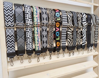 May include: A display of 16 different patterned black and white guitar straps with silver hardware. The straps are hanging on a wooden shelf.