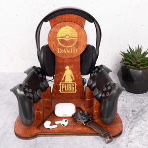 Può includere: Supporto in legno per cuffie con nome personalizzato "DAVID" e logo Pokemon. Ha anche due slot per i controller di gioco e un piccolo scomparto per altri accessori. Il supporto è marrone e ha un logo PUBG.