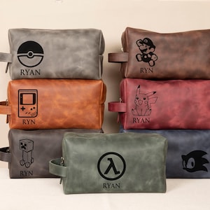 May include: Six different personalized leather toiletry bags with various gaming character designs. The bags are brown, gray, red, green, blue, and orange. Each bag has a different character design, including a Pokeball, a Game Boy, a Minecraft character, a Half-Life logo, a Sonic the Hedgehog character, and a Pokemon character. Each bag also has the name "Ryan" printed on it.
