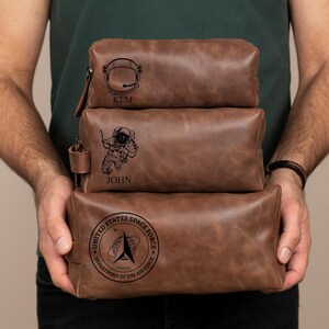 May include: Three brown leather zippered pouches with personalized engravings. The top pouch has a space helmet and the name "KIM". The middle pouch has an astronaut and the name "JOHN". The bottom pouch has the United States Space Force logo and the text "DEPARTMENT OF THE AIR FORCE".