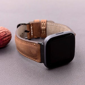 May include: A brown leather watch band with a dark gray smartwatch. The band has a distressed look and is stitched with white thread.