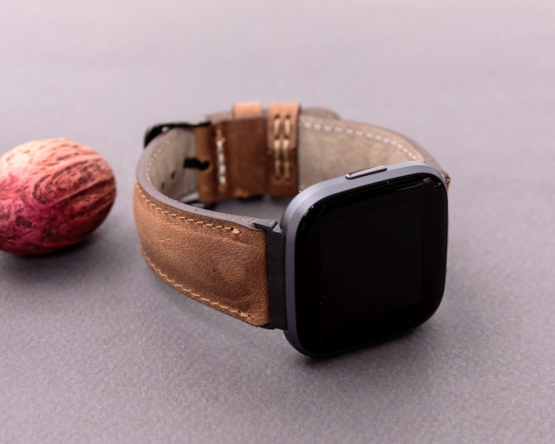 May include: A brown leather watch band with a dark gray smartwatch. The band has a distressed look and is stitched with white thread.